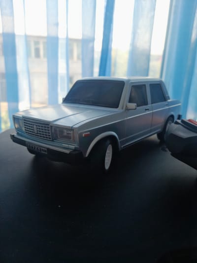The image shows a silver toy car, possibly a model of a Lada, placed on a dark surface. The car is facing towards the left of the frame and is slightly angled. The background consists of sheer blue curtains with sunlight filtering through them, creating vertical stripes of light and shadow. Behind the curtains, a blurry building facade is visible, suggesting an indoor setting near a window. The car has a license plate that reads "TT7ZY". A black object with a red element is partially visible in the lower right corner of the image, possibly a remote control. The lighting suggests it is daytime. There are no people or animals present in the image. The scene is static and does not depict any action. The location in Bayaut, Uzbekistan is not discernible from the image itself.