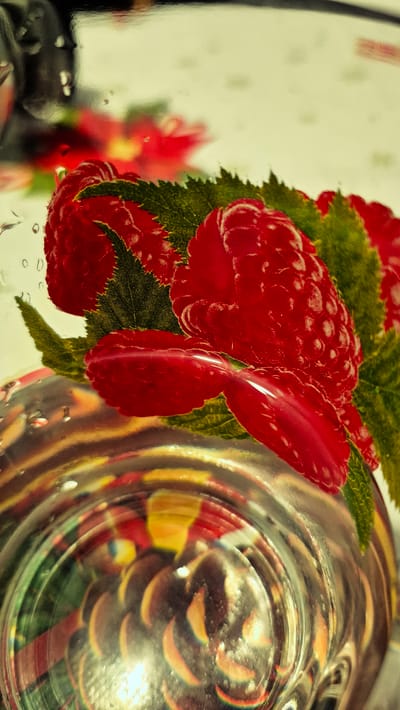 This is a close-up, vertically oriented, macro photograph of a printed image of raspberries and green leaves. The printed image is seen through a curved, transparent surface, possibly glass or plastic, which has water droplets on it and shows distorted reflections of other objects. The raspberries are red and appear textured, with visible seed-like imprints. The green leaves are serrated along their edges. The background of the printed image is a light cream color with faint red floral patterns. The transparent surface creates a refractive effect, showing concentric circular patterns and distorted colorful reflections from objects beneath it. The overall lighting is bright and somewhat diffused, with some areas of highlight and shadow. The photograph is taken from a low angle, looking upwards towards the printed image. There are no people or visible signs of activity. This image does not depict any location in Troițcoe, Moldova.