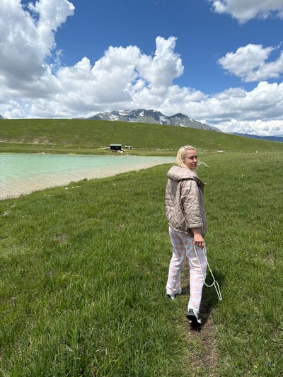 The image displays a wide landscape in Javorje, Montenegro, featuring a female individual walking on a dirt path through extensive green grasslands under a dynamic blue sky with large, white cumulus clouds.

In the foreground, a blonde woman is seen from behind, walking on a narrow, dark dirt path that meanders through dense green grass. She is turned to her right, looking directly over her shoulder towards the viewer with a neutral expression. Her blonde hair appears to be lightly wind-blown. She is dressed in a light brown or beige quilted jacket with a hood, featuring three-quarter length sleeves. Her lower attire consists of light-colored plaid pants, patterned with pink, white, and faint darker lines. She wears dark athletic shoes with white soles. In her right hand, she carries a white, flexible, rope-like object forming a loop.

To the left of the image, a light turquoise-colored lake with clear water is visible, bordered by a lighter-toned shoreline. Beyond the lake, a small, dark-colored wooden building is situated near the base of a verdant hill. Adjacent to the building, a white SUV is parked, and several dark-colored animals, possibly horses or cattle, are present. A yellow object, resembling a kayak, is partially submerged or beached at the lake's edge opposite the viewer.

The midground and background consist of gently rolling green hills and mountains. The distant mountain range exhibits patches of snow on its peaks and slopes under the bright blue sky. The overall scene depicts a serene, open natural environment with distinct geological and hydrological features.