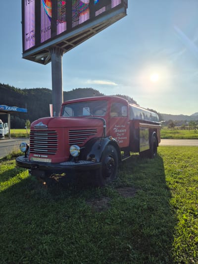 An old red and black fuel truck is parked on a grassy area next to a road. The truck has a large tank on the back and the words "Karl u. Anna Thrairer Dorf 36, 242 Niederndorf, 042 Niederndorf 2405" written on the side of the cab. In the background, there is a large billboard displaying colorful graphics and text that appears to be advertising. The sun is setting behind a hill, casting a warm glow on the scene. The location is identified as Niederndorf, Austria.