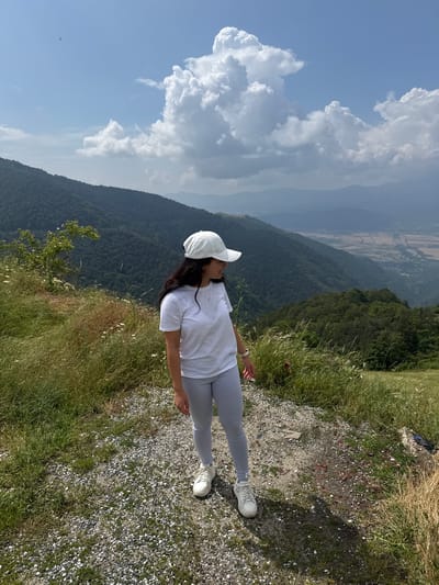In the city of Hristo Danovo, Bulgaria, a young woman stands on a gravel path at the edge of a mountain landscape. She is wearing a white t-shirt, light grey leggings, a white baseball cap, and white sneakers. Her dark hair is visible beneath the cap, and she is looking off to her right. The background features rolling green hills covered in trees, with a valley and distant mountains visible under a partly cloudy sky. The foreground consists of a gravel path with dry grass and some wildflowers growing along the sides.