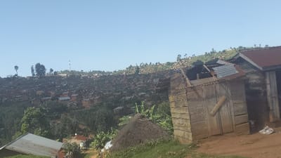The image shows a scene in a hillside slum in Rutshuru, Democratic Republic of Congo. The foreground contains a dilapidated wooden structure with a corrugated metal roof, and the hillside behind is densely packed with small, simple dwellings. The houses appear to be made of brick and wood. Trees and foliage are interspersed throughout the scene. The sky is clear, with the sun shining. No people are visible in the image.