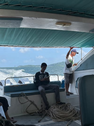 The image shows the interior of a boat traveling on the sea near Pak Nam, Thailand. Two men are visible. One man, wearing a black sweatshirt with a white graphic and grey pants, is sitting on a blue bench and looking at his phone. The other man, wearing a white t-shirt with a graphic, grey shorts, and a blue baseball cap, is standing and holding onto a railing on the right side of the boat. He is looking out towards the sea. Several boat engines are visible on the back of the boat, and a large coil of rope is on the floor in the foreground. The sea is choppy with white wake, and a green buoy and a distant coastline with buildings and greenery are visible in the background under a cloudy sky.
