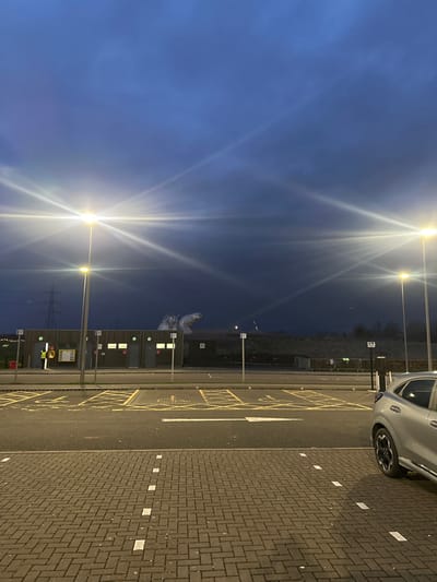 This image depicts an outdoor car park or service area at dusk or night, located in Falkirk, United Kingdom, with a view of The Kelpies sculptures. The sky is dark blue-grey and overcast.
In the foreground, the ground is paved with reddish-brown interlocking bricks, marked with white dashed lines. An asphalt section features yellow crosshatch "keep clear" markings and a white arrow indicating direction. The right side shows the partial passenger side and rear wheel of a silver or light grey Ford Puma SUV, parked next to an electric vehicle charging station.
The midground is dominated by multiple tall streetlights emitting bright, star-shaped lens flares, illuminating the scene. A row of low-rise utility buildings or public toilets, constructed with wooden paneling and white doors/windows, stands horizontally. One individual, wearing a high-visibility yellow vest, is visible near the leftmost building. Grey bollards or short posts are positioned along the edge of the parking area.
In the background, beyond the buildings, is an elevated, sparsely vegetated landform. Prominently situated in the distance on the left are two large, metallic, equine-head sculptures, identified as The Kelpies. Further left, a tall electricity pylon and power lines are visible against the horizon. No clear actions or interactions among people are discernible beyond the stationary individual.
