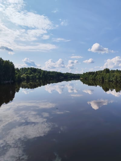 This is a landscape photograph of a wide, calm body of water, likely a river or lake, reflecting a bright blue sky filled with scattered white cumulus clouds. The shoreline on either side is densely lined with a variety of green trees, forming a thick forest. The reflection of the sky and the trees is mirrored almost perfectly on the glassy surface of the water. A small, dark object, possibly a buoy or marker, is visible in the distance on the right side of the water. The location is identified as Verkhnii Rubezh, Russia.