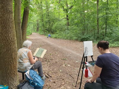This media file depicts two individuals engaged in an art activity outdoors in a forest setting, likely in Falkensee, Germany. The scene is dominated by lush green foliage and a dirt path winding through the trees, suggesting it is daytime and the weather is pleasant.

On the left side of the image, a person wearing a dark blue t-shirt is seated, facing an easel. The easel holds a white canvas with some charcoal or dark marks on it. A red water bottle and other art supplies are visible on a portable stand next to the person.

To the right, another individual, with grey hair, is also seated and focused on their artwork. They are wearing a light-colored shirt with a floral pattern. In front of them is a sketchpad or board with a drawing that appears to be of tree roots. A blue backpack rests behind them, along with a water bottle. The presence of a tripod-like structure on the left suggests it might be a camera tripod or part of an easel setup.

In the background, further down the path, a third person, possibly a child, is visible in the distance, wearing a blue and green outfit and riding what appears to be a scooter or a small bike. The overall atmosphere is one of quiet concentration and creative engagement within a natural environment. No text is visible in the image.
