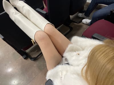 A person is seated, wearing a white faux fur coat. Their legs are crossed, clad in tan tights and white knee-high boots. They are seated in a theater with red upholstered seats. The floor is a marbled stone. Another person's legs can be seen in the lower left of the frame.
