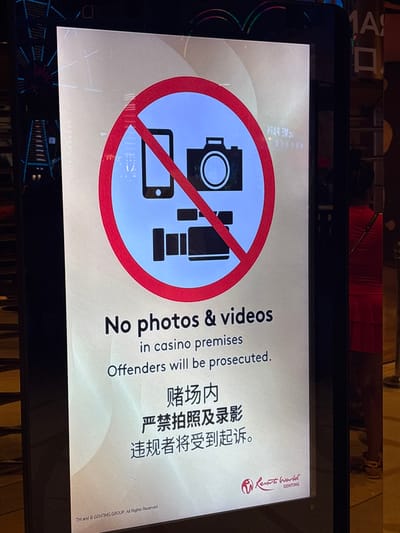 The image shows a sign in Genting Highlands, Malaysia, displaying a prohibition symbol over icons of a camera, smartphone, and video camera, indicating that photos and videos are not allowed on casino premises. The text reinforces this message in English and Chinese, stating that offenders will be prosecuted. The Resorts World Genting logo is visible in the top right corner.