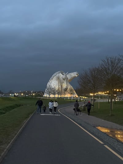 The image depicts an outdoor scene at dusk or early evening, centered on The Kelpies sculpture in The Helix park, Falkirk, United Kingdom. The colossal, illuminated, silvery metallic sculpture of two horse heads dominates the background, rising against a dark, overcast sky.
In the foreground, a dark asphalt path curves from the lower left towards the upper right. A family group of four individuals walks towards the viewer on the left side of the path: an adult male in dark attire, a small child in dark clothing, and two adult females in light-colored jackets. Further along the path, moving away from the viewer on the right side, an adult male in dark clothing walks a leashed, dark-colored dog. To his right, another adult, wearing a dark jacket and a red hat, walks with arms slightly extended.
The path features a white solid line and a dashed white line separating it from a gravel shoulder on the right, which shows areas of reflective wetness. A grassy verge runs along the left side of the path. Bare deciduous trees are present throughout the scene; some are visible between the path and The Kelpies, while others line the right side of the path, illuminated by strings of warm-toned outdoor lights strung between them. Distant vehicle lights or streetlights are visible on the far left horizon. The overall lighting suggests low ambient light conditions, with primary illumination originating from the sculpture's base and the decorative string lights.
