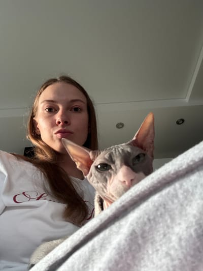 A young woman with long, brown hair is in the foreground, looking directly at the camera with a neutral expression. She is wearing a white t-shirt with red lettering. Partially obscuring the lower right side of the image is a Sphynx cat, whose head is visible, looking towards the left. The cat has grey and pink skin and large ears. The background shows a textured ceiling with recessed lighting fixtures. The scene appears to be indoors, possibly in a residential setting in Novaya Ivanovka, Russia.