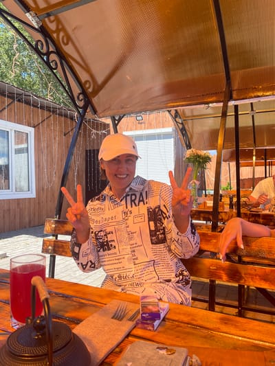 A woman wearing a white baseball cap and a shirt with a newspaper print sits at a wooden table outdoors under a translucent amber canopy. She is smiling and holding up two fingers in a peace sign with each hand. In front of her is a glass of red liquid, a black teapot, a folded napkin with a fork, and a pack of cigarettes. The background shows a wooden building and trees. The scene is likely a cafe or restaurant in Belozersk, Russia.