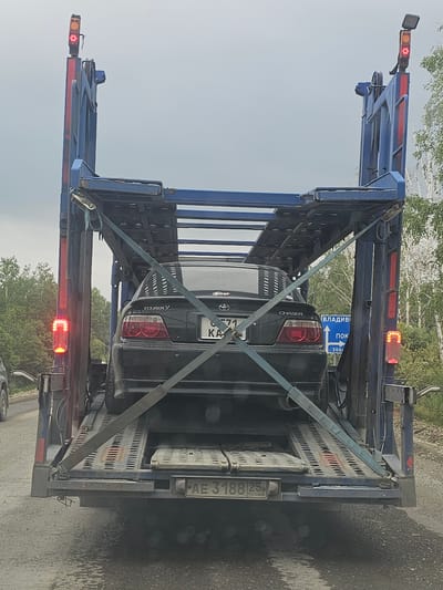 An overhead view of a black Toyota Chaser Tourer V being transported on a car carrier trailer is captured on a cloudy day. The vehicle, with license plate "K 71 KA," is secured to the trailer with blue straps. The trailer itself is blue and has red brake lights visible on its rear. A blue road sign with white text and arrows is attached to the right side of the trailer, indicating directions to "ВЛАДИВ" and "ПОК," with a distance of "300." The trailer's license plate reads "AE 3188 25 RUS." The environment appears to be a rural roadside with trees and a gray, wet road surface, suggesting it might be located near Lesopil'noe, Russia.