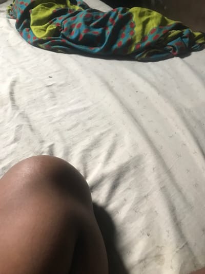 The image depicts a close-up, top-down view of an indoor setting in Isasi, Nigeria, focusing on a bed. In the lower-left quadrant, a portion of a human leg, specifically the upper thigh and knee area, with dark skin tone, is visible. The leg rests on a wrinkled white or light-colored bed sheet, which exhibits subtle, small patterned impressions (resembling stars) and minor scattered debris or discolorations. In the upper half of the frame, a rolled or folded piece of fabric, possibly a blanket or wrapper, is present. This fabric displays a vibrant pattern consisting of a teal or blue base with red polka dots, interspersed with sections of lime green featuring darker, abstract patterns. The lighting suggests an artificial source, casting soft shadows.