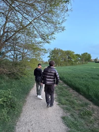The image, taken in Gometz-le-Châtel, France, shows two men walking along a gravel or dirt path through a natural landscape. The photo is vertically oriented, with the path leading away from the viewer towards the upper right.
The subjects are two men, viewed from behind. The man in the foreground wears a dark, possibly black, puffy jacket and dark pants, with short, dark hair. The second man, positioned slightly behind and to the left of the first, is dressed in a dark jacket, light-colored (possibly olive green or grey) pants, and white athletic shoes.
The scene is dominated by a clear, bright blue sky at the top. To the left of the path, a dense arrangement of trees with green foliage suggests a wooded area or tree line. To the right of the path, a wide expanse of vibrant green grass stretches out, resembling a field or park. In the far background, faint structures, likely buildings or houses, are visible on the horizon, hinting at a nearby settlement.
The activity depicted is a leisurely walk. The bright, clear sky indicates pleasant weather, and the overall lighting suggests it is daytime, possibly late afternoon or early evening. No text is visible in the image. The setting portrays a serene, semi-rural environment.