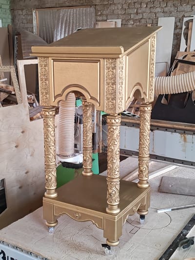 A gold-colored, ornate wooden lectern stands on a light-colored surface with intricate carvings. The lectern has a slanted top, decorative carvings on its sides, and four carved columns supporting the top section. The base of the lectern has a shelf and is supported by four round feet, two of which have small casters. The background appears to be a workshop or storage area, with stacks of wood, brick walls, and industrial equipment. The overall lighting is natural, suggesting daylight. The specific location of Ичетовкины, Russia, is not determinable from the visual information provided.