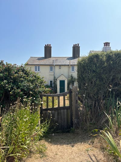 The image depicts a quaint, pale yellow cottage with a gray tiled roof, set against a clear blue sky. The cottage features multiple chimneys, white-framed windows, and a dark green door. In the foreground, a rustic wooden gate stands slightly ajar, revealing a pathway leading towards the house. Lush green bushes and tall grasses with small white flowers flank the gate and path, suggesting a garden setting. The overall impression is one of peaceful rural charm. This cottage is located in Seaford, United Kingdom.