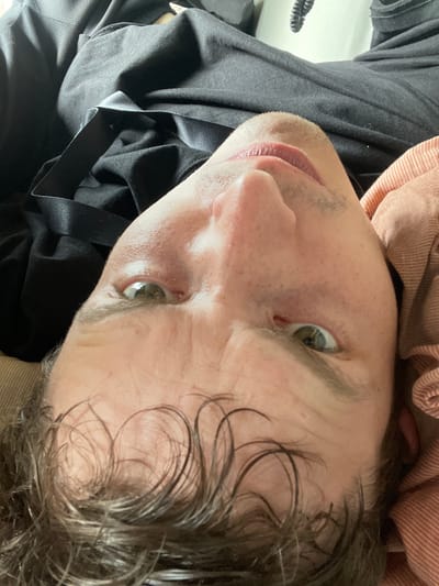 The image is a close-up, upside-down, selfie-style shot of a person's face. The person has light skin, brown hair with some lighter strands, and green eyes. There are wrinkles on the forehead and around the eyes. The person is wearing dark clothing, possibly a t-shirt, and a pink and white striped garment is visible on the right side. The background is blurred and appears to be indoors, with a hint of a white surface and a black cord-like object in the upper right corner. There is no indication of the location in Edewecht, Germany.