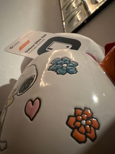 The image shows a close-up of a white, glossy ceramic mug, possibly a souvenir, decorated with cartoon-like illustrations. On the mug's surface, there are several embossed and painted designs: a stylized Snoopy character's eye, a pink heart, a blue flower with six petals, and an orange flower with six petals. A small, white tag is partially visible, angled over the top of the mug, with text that reads "Gideon Bible App" and "Find Help. Find Hope." The background is a neutral, light-colored surface, with a reflective, metallic-like object or surface visible in the upper portion of the image. The lighting is somewhat dim, creating highlights and shadows on the ceramic surface. The location in Lynnwood, United States is not discernible from the visual information provided.