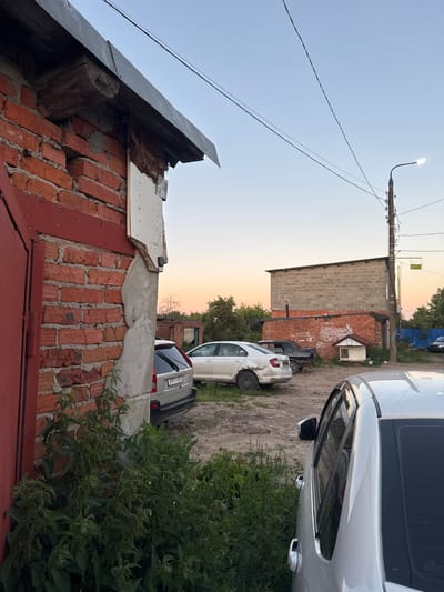 The image captures an outdoor scene, likely in Боголюбово, Russia, during what appears to be late afternoon or early evening, given the warm, soft light of the setting sun. The primary subjects are several cars parked on a dirt or gravel lot. One prominent silver car is in the foreground, its side and rear visible. Another white car is parked behind it. In the background, a brick building with a lighter colored block-built section is visible, with some signs of disrepair, particularly around the roofline. Trees and greenery are present, suggesting a somewhat rural or suburban environment. Overhead, a street lamp is illuminated, and electrical wires stretch across the sky. A portion of a red-tiled roof or wall is in the lower part of the frame, adding to the impression of a residential or industrial area. There is no clear activity or event taking place; the cars are parked, and the scene appears static. Notable details include the construction materials of the buildings and the condition of the foreground roof, which shows exposed wood and crumbling plaster. The sky is clear, indicating fair weather. A yellow sign with text is visible on the building in the background, but the text is not legible. A license plate on the silver car shows the text "X977 X069".