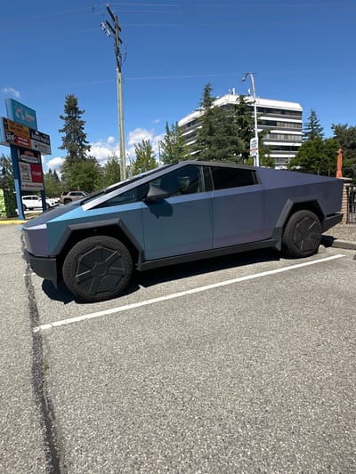 A Tesla Cybertruck, with a matte blue-purple iridescent wrap, is parked in a lot in Port Coquitlam, Canada. The vehicle is facing left and is angled slightly towards the viewer. It is parked in a designated parking spot, indicated by a white line on the asphalt. In the background, there are trees, a light pole with wires, a utility pole, and a multi-story building. Signs are visible on the left side of the image, including one that reads "FOR LEASE" and another with "RE/MAX". The sky is clear and blue with some scattered clouds. The overall scene depicts an urban or suburban environment on a sunny day.