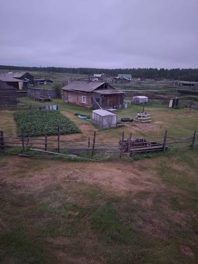 The image depicts a rural village scene in the city of Kyuelyany, Russia, under an overcast sky. The foreground features a wooden fence and dry, patchy grass. Beyond the fence, a large, well-tended garden bed is visible, bordered by more dry grass. To the right of the garden are several structures, including a wooden house with a window, a greenhouse, and what appears to be a woodpile. In the background, more wooden houses, sheds, and agricultural outbuildings are scattered across the landscape, with a line of trees forming the horizon. The overall impression is one of a remote, self-sufficient settlement.