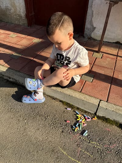 In Hadzhidimitrovo, Bulgaria, a young child, identified as a boy, is depicted outdoors in a sunlit environment. The child is seated on the edge of a raised, reddish-brown tiled pavement, which features small, lighter square inserts and a textured surface. He is positioned facing slightly left, with his head tilted downward, attentively observing a scattered collection of sidewalk chalk on the asphalt surface below.
The child has short, dark brown hair with a distinct shaved line design on the right side of his head. He wears a white short-sleeved t-shirt with a dark graphic print on the front, partially visible as "STAR," possibly related to Star Wars. Below, he wears black shorts. His feet are clad in white socks, one displaying a brown leaf-like pattern and the other a grey patterned design, and light blue Crocs-style clogs. The clogs are adorned with multiple colorful character charms, including a green figure, a red Thor's hammer, a white/blue robot, and a red mushroom.
A grey concrete curb separates the tiled pavement from the dark asphalt surface. On the asphalt, approximately 20-30 pieces of chalk in various colors (yellow, orange, pink, light blue, green, purple) are distributed in a pile directly in front of the child. Faint yellow and pink chalk lines are visible on the asphalt, suggesting prior drawing activity. Small patches of green moss are present along the base of the curb on the asphalt side.
Strong, natural sunlight from the left casts pronounced shadows across the tiled pavement, indicating a low sun angle. In the background, a textured light-colored wall is visible to the left, and a dark reddish-brown door or gate with a metal frame is partially visible to the right. The child's hands are resting, with his right hand on his right knee/thigh and his left hand near his left ankle, indicating a pause in activity or readiness to engage with the chalk.