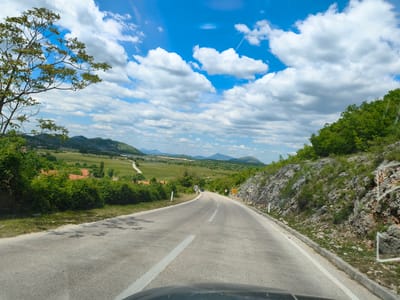 The image shows a road winding through a rural landscape near Grab, Bosnia and Herzegovina. The road is paved and has a white line dividing the lanes. The sky is blue with scattered white clouds. Green vegetation covers the hills and mountains in the background. A tree with sparse foliage stands to the left of the road. A rocky slope with sparse vegetation is visible on the right side of the road. There is a vehicle in the distance on the road. The image appears to have been taken from inside a vehicle, as part of the dashboard is visible at the bottom.