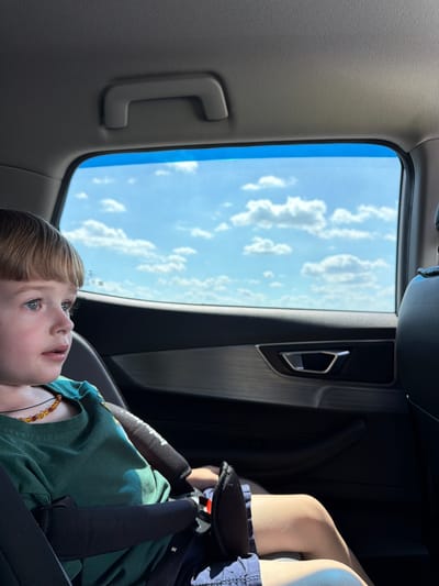 The image shows a young child with blond hair and blue eyes looking up at the sky from inside a vehicle. The child is wearing a green t-shirt and appears to be in a car seat, with safety straps visible. The setting is the interior of a car, with a window showing a bright blue sky with scattered white clouds. The sun appears to be shining, suggesting it is daytime. A grab handle is visible on the left side of the interior, and a door handle with metallic trim is visible on the right. The overall mood is peaceful, with the child seemingly captivated by the view outside. There is no visible text in the image. The location context is Zapadny, Russia.