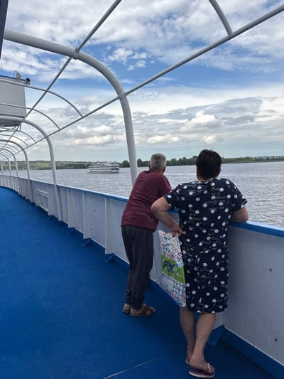 Two people are on the deck of a boat, looking out at the water. The person on the left is a man with graying hair, wearing a maroon shirt and dark pants. The person on the right is a woman with dark hair, wearing a dark blue dress with white polka dots and a patterned tote bag. The boat has a blue deck and a white railing. In the distance, a large white boat is visible on the water. The sky is cloudy. The location is the city of Bet'ki, Russia, on the Volga River.