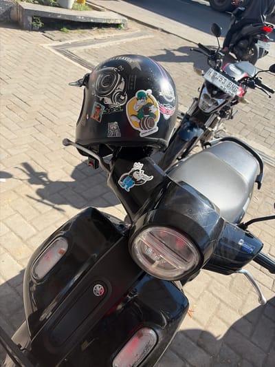 The image shows a close-up view of a black scooter parked on a paved walkway. The scooter's handlebar and front section are prominent, with a helmet resting on the handlebar. The helmet is black and adorned with several stickers, including one with a cartoon character holding a barrel and another depicting a stylized engine. A license plate, "H 2453 BNG," is visible on the front of another motorcycle parked behind the scooter. In the background, a person is seated on the rear motorcycle. The scene appears to be outdoors in an urban setting, with sunlight casting shadows on the ground. The location is identified as Jipang, Indonesia.