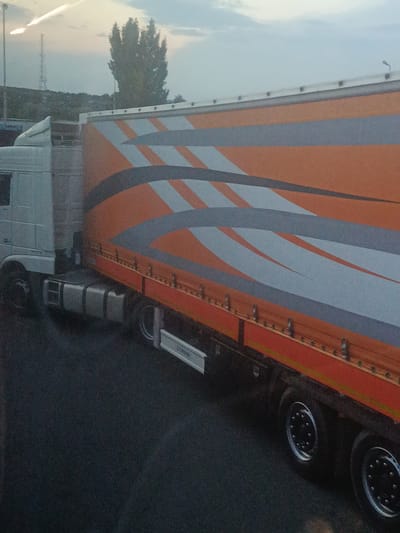 The image shows a white truck with an orange and grey striped trailer parked on a dark asphalt surface. The trailer has a series of metal latches along its lower edge. The truck cab is on the left side of the image, and the trailer extends to the right. In the background, there is a hazy sky with some clouds, a distant treeline, and a tall tower or antenna. The overall lighting suggests it might be dusk or dawn. The scene appears to be an outdoor parking area, possibly a rest stop or industrial zone. There are no people visible in the image, and no discernable action is taking place. The location is identified as Oancea, Romania.