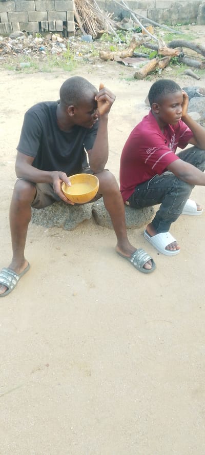 This media file captures two young Black males sitting outdoors in what appears to be Doka-Tofa, Nigeria. The setting is sandy with scattered rocks and some debris in the background, including construction materials like cinder blocks and dried vegetation. The lighting suggests it is daytime, possibly late morning or early afternoon, with soft, diffused light.
The subject on the left, wearing a black t-shirt and brown shorts, is seated on rocks and holding a hollowed-out gourd filled with a light-colored liquid, possibly a beverage. His gaze is directed to the side, and his hand is touching his forehead, conveying a pensive or perhaps weary emotion. He is wearing dark, striped slide sandals.
The subject on the right, dressed in a maroon polo shirt with subtle white embroidered designs and dark, distressed jeans, is also seated on rocks. He is looking to the side with a relaxed posture, his hand resting on his knee. He is wearing white slide sandals.
The overall atmosphere is relaxed and informal. There is no visible text in the image. The scene depicts a quiet moment shared between two individuals in an open, somewhat unkempt outdoor environment.