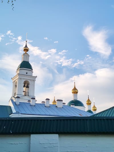 This media file captures an outdoor scene featuring religious architecture under a bright, partly cloudy sky. The primary subjects are several domed structures, characteristic of Russian Orthodox churches, with a prominent white bell tower featuring a green dome and a golden cupola topped with a cross. Smaller golden domes, also adorned with crosses, are visible in the background, some with green bases. The buildings are partially obscured by layers of dark green and blue corrugated metal roofs in the foreground.
The setting appears to be Vasil'ki, Russia, a detail suggested by the architectural style. The time of day is likely late afternoon or early evening, as indicated by the soft, warm lighting illuminating the golden domes and the contrast with the blue sky and white clouds. The weather is fair, with scattered clouds creating dynamic patterns against the clear sky.
No people are visible in the frame. The activity suggested is purely architectural observation. Notable details include the intricate designs of the golden domes and crosses, the architectural details of the bell tower, and the contrast between the traditional religious structures and the more modern roofing materials. The overall mood is serene and somewhat majestic, evoking a sense of spiritual presence and historical significance. There is no visible text in the image.
