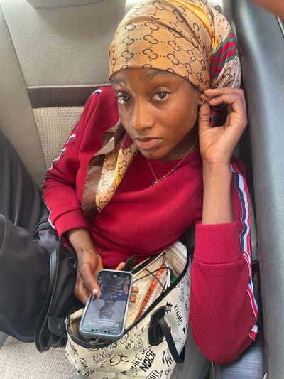 This media file shows a person sitting in a vehicle, likely a car, in Bassa, Nigeria. The person is wearing a red sweatshirt with striped sleeves and a headscarf patterned with the Gucci logo and green, red, and yellow stripes. They are holding a smartphone in their right hand, which displays a video of a person. The person in the image is touching their ear, possibly adjusting an earring. A black purse and a patterned tote bag are visible next to them. The tote bag has text and drawings on it, including the word "YINMEI" and other illegible writing. The person's expression appears neutral or slightly contemplative. The seating in the vehicle appears to be made of grey fabric. The overall scene suggests a casual moment, perhaps during a commute or a break while traveling. The lighting suggests it is daytime.