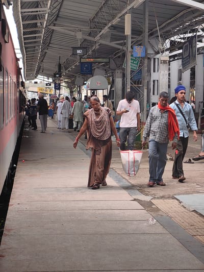 The image captures a bustling scene at Khandwa Railway Station in India. In the foreground, a woman in a brown salwar kameez and a patterned scarf walks towards the viewer, her right arm extended outwards. Behind her, a man in a checkered shirt and orange scarf carries a large white and red plastic bag. To his right, a young man wearing a blue turban and a light blue shirt walks with a crossbody bag. Further back, several people are visible on the platform near a train on the left, with some appearing to be boarding or disembarking. The platform is covered by a large, sloped metal roof supported by metal beams. Signage indicating platform numbers and directions is visible overhead. The environment suggests a typical Indian railway station with concrete platforms and a busy atmosphere.