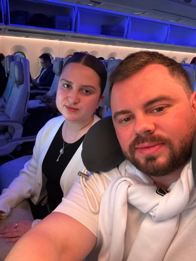 A selfie on an airplane captures a fair-skinned couple. The man has a beard and wears a white hoodie with a black travel pillow, leaning into the shot from above. The woman, with dark hair pulled back, wears a black top, a white cardigan, and a heart-shaped key necklace. Airplane seats and blue-toned lighting are visible in the background, suggesting an airplane interior.