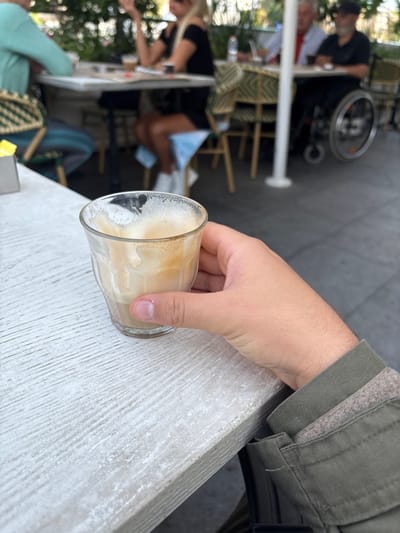 The image presents a first-person perspective at an outdoor cafe in Nahariya, Israel. In the foreground, a right hand with short fingernails holds a clear glass containing a partially consumed light brown beverage with white foam and residue clinging to the interior. The hand rests on a light-colored, textured table surface, resembling distressed wood. The individual wears a green-grey long-sleeved jacket with a visible lighter grey/beige lining at the cuff.
The background, out of focus, reveals other patrons seated at similar tables with woven chairs. To the left, a person in a light blue/green top is partially visible. Further back, a woman with long blonde hair wears a black top, with a blue face mask draped over her knee. To her right, an older man with grey hair wears a light-colored shirt with a red element. Adjacent to him, a man with a dark cap and beard is seated in a black manual wheelchair. The ground is a dark grey paved surface. A white vertical pillar is present, and green foliage is visible in the distant background, suggesting an outdoor dining area.