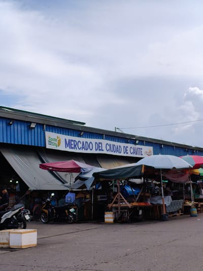 This image captures a street market scene in Cavite City, Philippines. The main subject is a row of market stalls under a large blue building with a metal corrugated facade. A prominent banner across the building reads "Cavite City MERCADO DEL CIUDAD DE CAVITE" in blue lettering.

The market stalls are covered with various tarps and awnings, some brightly colored like a red umbrella, and others in muted tones of gray and green. Beneath the awnings, goods are displayed, including fruits and vegetables arranged on tables and crates. Several motorcycles are parked in front of the stalls, indicating transportation for vendors or customers. A few people are visible in the market, though their features are not distinct enough to describe their activity or emotions, they appear to be engaged in market operations. One person, a woman with blonde hair, stands near a motorcycle on the left side of the image.

The setting appears to be an outdoor marketplace, likely during daylight hours given the overall illumination. The sky is partly cloudy, suggesting a moderate weather condition. The ground is paved, likely asphalt, with some scattered debris or small items. The presence of wooden crates and pallets used for displaying goods are notable details. The overall atmosphere suggests a busy, functional market environment.