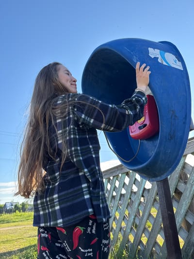 A young female, positioned on the left side of a blue outdoor payphone booth, is depicted in Ust'-Silayka, Russia. She is seen from the right side, facing towards the interior of the booth with a slight smile. Her long, light brown hair extends down her back and right shoulder. She wears a dark blue and green plaid fleece shirt and black pants adorned with red chili pepper motifs and the German text "Salz und Pfeffer" (salt and pepper). Her right hand, bearing a light-colored bracelet, reaches into the blue booth towards a red payphone unit. The blue booth features a partially torn sticker with Cyrillic text "АЛЬНЫЕ СВЯЗИ" and the "Ростелеком" logo. Behind the woman and the booth, a weathered wooden lattice fence with peeling light blue paint is visible. The ground is covered with green grass, and in the distant background, several indistinct buildings and power lines are present under a clear blue sky. The scene is outdoors during daylight hours.
