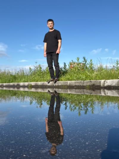 This is a vertically oriented, full shot of a man standing outdoors reflected in water. The man, wearing a black t-shirt and dark pants, appears to be posing for the camera. He is positioned on the right side of the image. The reflection shows him mirrored in the water, creating a symmetrical composition. The scene is set by a narrow, raised concrete pathway running vertically down the center of the image, separating the water on the left from lush green vegetation and a bright blue sky with scattered clouds on the right. The reflection in the water shows the sky, vegetation, and the man's mirrored image, with the water's surface appearing somewhat textured with small white specks. The lighting suggests it is daytime, likely a sunny day, given the clear blue sky. The overall impression is one of a playful or artistic interpretation of a common outdoor scene, utilizing the reflective quality of the water to create a striking visual effect. No text is visible in the image.