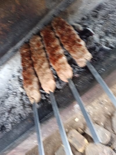 An overhead view captures four elongated ground meat kebabs, each impaled on a flat metal skewer, positioned over a bed of glowing embers and grey ash within a dark-colored metal brazier or grill structure. The kebabs exhibit cooked surfaces ranging from light brown to reddish-brown, with some localized charring, indicating active thermal preparation. The metal skewers extend towards the lower right, where additional dark, irregular objects, consistent with unburned charcoal or wood, are visible. The upper background is a dark, indistinct surface. No human figures or direct interactions are visible in the scene. This event is occurring in Kasri Arifan, Uzbekistan.