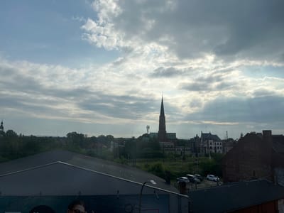 This is an eye-level, medium shot photograph taken from a high vantage point, looking out over a city landscape in Mysłowice, Poland. The sky dominates the upper half of the frame, filled with a mix of bright sunlight filtering through scattered clouds and darker, heavy cloud formations. In the mid-ground, a tall, slender church spire with a cross at its peak rises above other buildings. Adjacent to the church is a red brick building with a green roof. Further to the left, a dark green church tower is visible, partially obscured by trees. Several other buildings, including some with ornate architectural details and pitched roofs, are scattered across the scene. A cell tower is also present in the distance. The foreground is occupied by the dark, sloped roof of a modern building. Below this roof, a large billboard featuring a woman's face is partially visible. In the lower right of the frame, the corner of a dark brick building is seen. A line of parked cars, including a white hatchback and several darker sedans, are visible on a paved area near the buildings in the mid-ground. Railway tracks and overhead lines are also present throughout the scene, suggesting an urban or industrial area. The overall impression is of a somewhat overcast day with dramatic cloud formations, set against a backdrop of a Polish city.