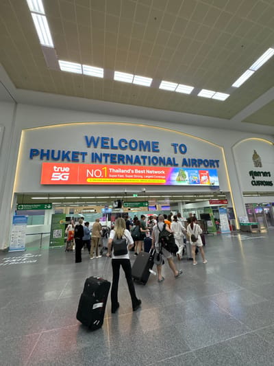 This image depicts the interior of the arrival area at Phuket International Airport, located in ตำบล ไม้ขาว, Thailand. Prominently displayed at the center is a large arched sign reading "WELCOME TO PHUKET INTERNATIONAL AIRPORT" in blue lettering. Below this sign, an illuminated LED screen displays a "true 5G" advertisement with the text "NO.1 Thailand's Best Network" and images of tourist destinations.
The floor consists of large grey tiles. The ceiling features a grid pattern with numerous rectangular fluorescent light panels providing bright illumination.
In the foreground, a person with light-colored hair, viewed from behind, wears a white t-shirt, black pants, black boots, and a black backpack. This individual is pulling two black rolling suitcases.
Approximately 20-30 individuals, predominantly travelers, are present in the midground and background, moving towards or through an area marked with directional signage. On the left, a green sign indicates "Nothing to Declare" with a downward arrow and a QR code poster. On the far right, a "CUSTOMS" sign in English and Thai script is visible above a processing area. Most individuals are observed walking, many pulling various types of luggage including rolling suitcases and carrying backpacks. No direct interactions between individuals are discernible.