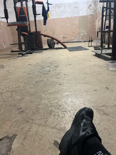 The image shows the interior of what appears to be a home gym. A black athletic shoe with black socks is visible in the upper right corner. Exercise equipment, including a multi-gym apparatus with orange and black upholstery, is in the lower left of the frame. A barbell with weights sits on a metal stand along the back wall, to the left. The walls are peeling and unfinished, with a distinct line between the patched and unpatched sections. The floor is a stained, light-colored surface. The image provides no indication of the location in Kalchevo, Bulgaria.