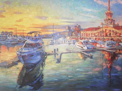 The painting depicts a harbor scene at sunset with several yachts docked. In the foreground, a large blue yacht is prominently featured, casting a reflection on the water. Several other yachts of varying sizes are visible in the middle ground, also docked. In the background, there is a city skyline with buildings and a tall tower, possibly a lighthouse or clock tower. The sky is filled with warm, sunset colors, including yellow, orange, and pink. The water reflects the colors of the sky and the surrounding structures. The overall impression is one of a peaceful and picturesque evening in a coastal city. The painting is done in an impressionistic style with visible brushstrokes. There is no indication that this scene is located in Gülnar, Turkey.