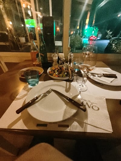 The scene depicts a dining table inside "No Pizzeria" located at Blvd. Gjorce Petrov 58, Gevgelija, North Macedonia, at night. The wooden table is set with placemats. In the foreground, a white ceramic plate holds a fork and knife with wooden handles, positioned diagonally. A stemmed wine glass containing rosé wine is present. To its left, a small light blue textured glass, a white bowl filled with a mixed salad, salt and pepper grinders, and a small clear glass carafe are arranged. Two bottles, one clear with a green label (partially obscured) and one amber-colored, stand behind these items. A second partially visible white ceramic plate with cutlery is positioned to the right. A white napkin with "Caffe Pizzeria No" text is visible under the right-side plate. Through a large window in the background, a nighttime street scene is visible, illuminated by distant green lights and featuring a red vehicle with illuminated taillights. Reflections of the interior lights are evident on the window glass. A person's bare leg is visible beneath the table in the bottom center.