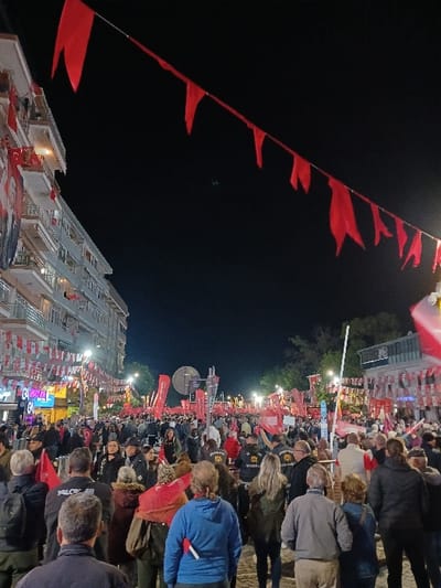 The image shows a large crowd of people gathered on a street in Silivri, Türkiye. The crowd is largely comprised of adult males and females, many wearing traditional Turkish clothing. The majority of people are standing and observing. Some individuals are speaking with others. There are red flags and banners strung across the street, and buildings line the streets. The scene is lit by street lamps and illuminated signs. The image suggests a lively, festive atmosphere. It is a night scene and the sky is dark.
