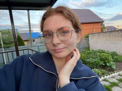 A young woman is depicted in a close-up, headshot-style image, appearing to be a selfie. She has light brown hair, which is slightly disheveled around her face, and numerous freckles are visible across her nose, cheeks, and forehead. She wears large, rectangular-framed eyeglasses, through which her light-colored eyes are visible, looking directly at the viewer. Her lips are slightly parted.
She is dressed in a dark blue zipped jacket or hoodie, with the silver zipper prominent on the front. Her right hand, adorned with a simple golden ring on her ring finger, is resting near her neck, with her left hand likely beneath it, out of view.
The background suggests an outdoor, possibly suburban or rural setting. Directly behind her, to the viewer's left, is a manicured garden area with various green plants and a pathway bordered by white stones. Further back, a light-colored brick wall and a wooden fence are visible. To the right, various building structures with corrugated metal roofs (brown and blue) are seen, alongside utility poles and scattered trees and greenery. The sky, featuring soft clouds and hues, indicates late afternoon or early evening. The overall location is Novaya Tura, Russia. No visible text is present in the image.