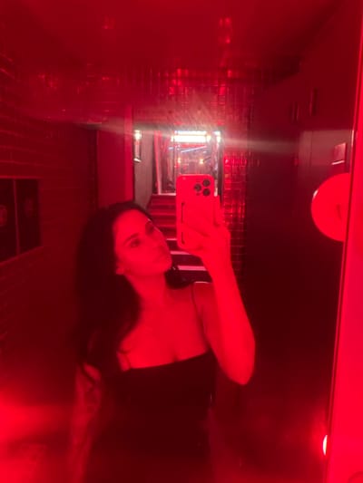 A young adult female is depicted from the mid-torso upwards, capturing a mirror selfie within a vividly red-lit indoor environment. She has dark hair, a neutral facial expression, and wears a black, thin-strapped garment. A tattoo is visible on her left arm. She holds a red smartphone, featuring three rear camera lenses, in her right hand, positioned to photograph her reflection. The entire scene is bathed in intense red light, casting a monochromatic hue over all surfaces. The reflective mirror surface reveals a narrow corridor or hallway behind the subject, which includes a visible staircase. The walls within this reflected space are constructed from dark, reflective materials, incorporating sections of small, glossy square tiles and larger, smooth panels. On the right side of the reflected background, a circular, illuminated fixture is present on the wall, while two dark rectangular panels with subtle emblems are visible on the left. The scene suggests an interior space consistent with a commercial or entertainment establishment in an urban setting. No specific identifying markers for 강남구, South Korea are discernible in the image.