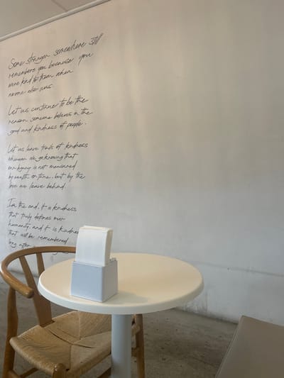 A white wall displays a handwritten black text about kindness and legacy. The words express that a stranger might remember you for your kindness and urge the reader to continue showing kindness. It also says that a legacy is not about wealth but about the love left behind. Furthermore, the text defines kindness as humanity and states it will be remembered. Next to the wall, there is a white table with a box of tissues on top of it and a wooden chair with a woven seat.